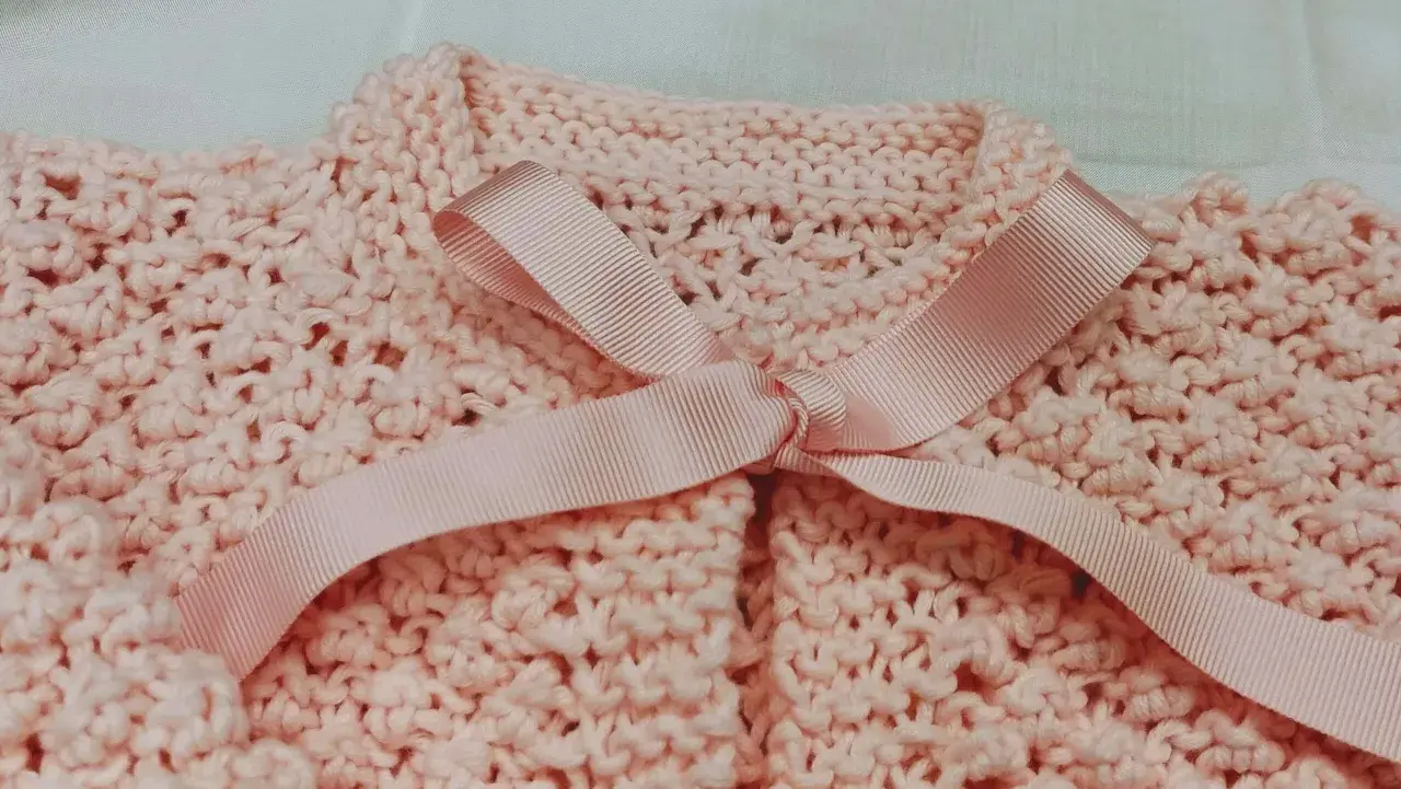 Dettaglio di un golfino bimba ai ferri, rosa pastello, con fiocco in raso. Spiegazioni per realizzarlo.