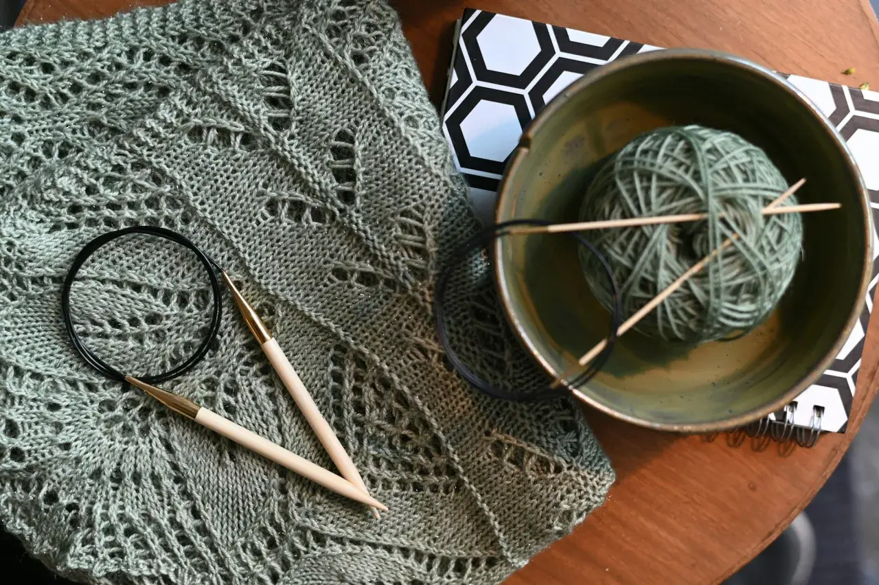 Lavori con lana sfumata: un maglione traforato, ferri da maglia, un gomitolo in una ciotola e un quaderno con motivo esagonale.