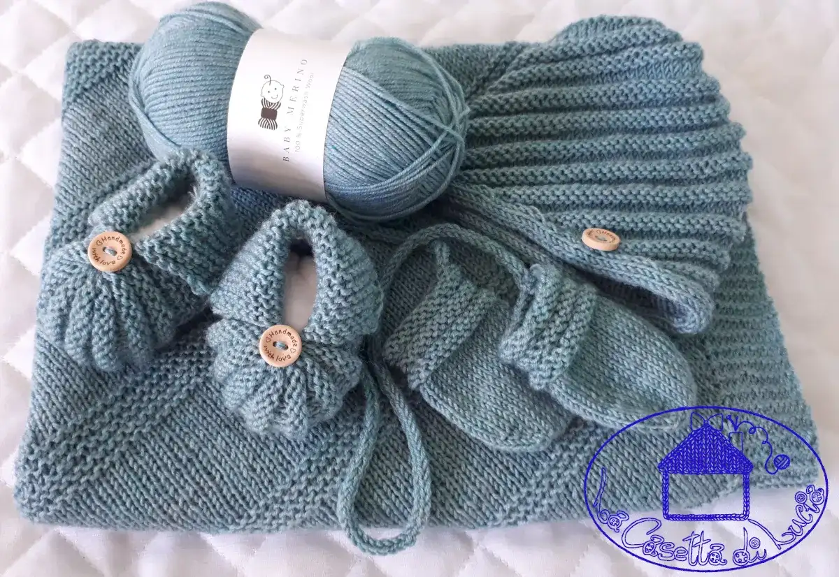 Un set per neonato ai ferri: copertina, scarpine e cappellino in lana merino azzurra, con bottoncini in legno.