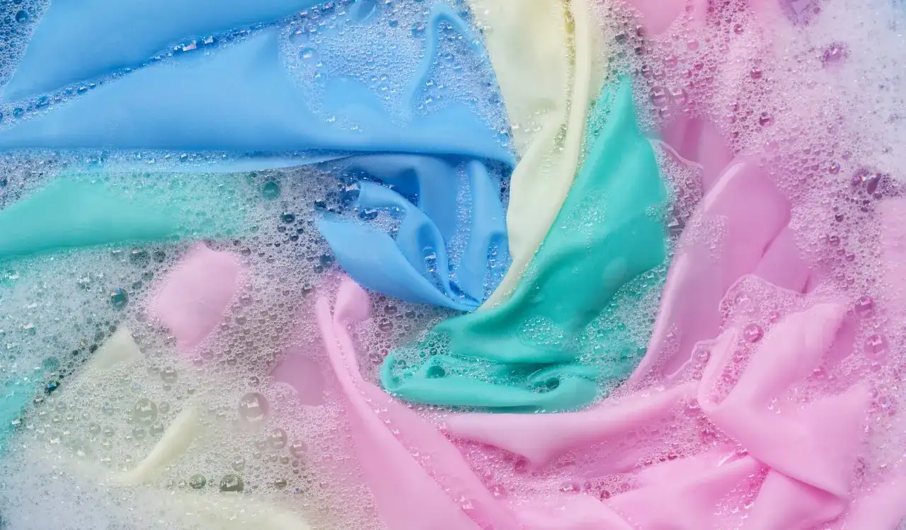 Tessuti color pastello, tra cui un **acetato tessuto** azzurro, si muovono tra bolle di sapone in un lavaggio delicato.