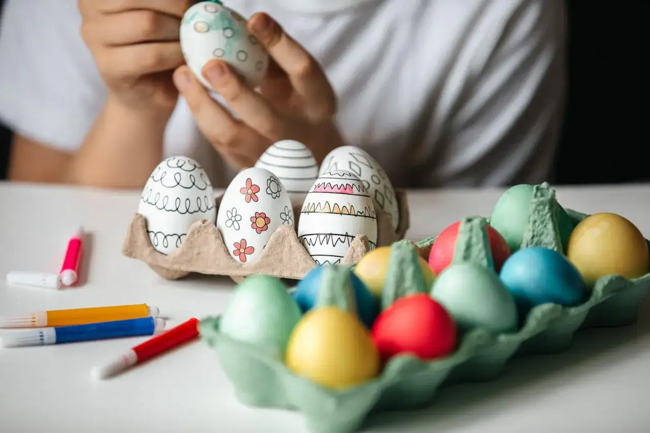 Bambino decora uova con pennarelli, creando disegni colorati. Sul tavolo, uova gi&agrave; dipinte e pennarelli pronti per i prossimi colori per uova di Pasqua.