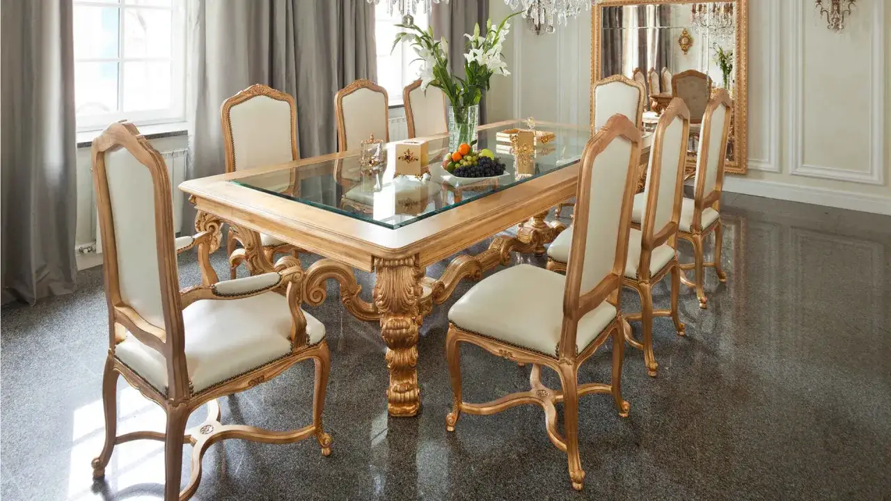 Tavolo da pranzo in legno dorato con piano in vetro, circondato da sedie imbottite. Un centrotavola con fiori e frutta aggiunge un tocco di colore a questo ambiente elegante, dove ogni dettaglio &egrave; un prezioso materiale.