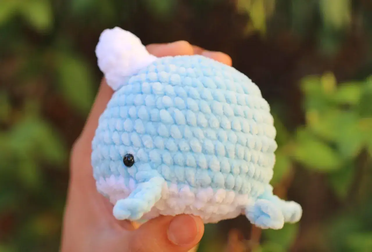 Balena azzurra fatta a mano con filati amigurumi, tenuta in mano su sfondo verde sfocato.