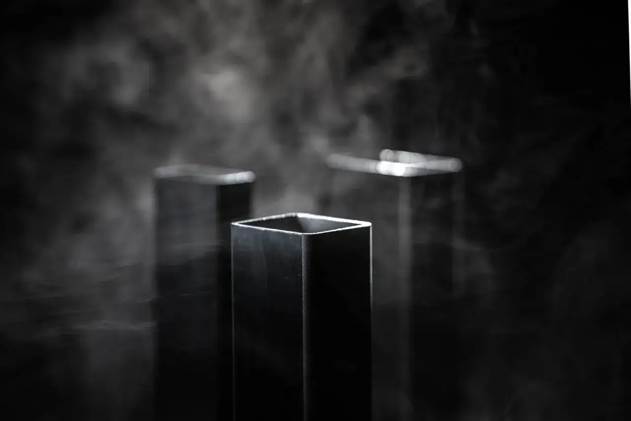 Profili quadrati in acciaio inossidabile diventano neri, avvolti da un fumo etereo su sfondo scuro.