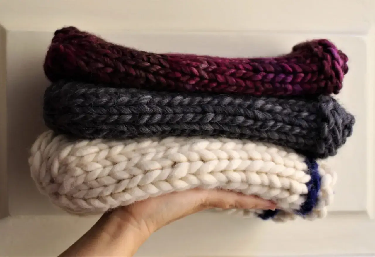 Mano che sorregge tre morbidi cappelli fatti a mano con lana grossa: bordeaux, grigio antracite e bianco panna con un dettaglio blu.
