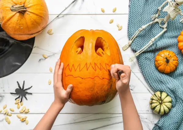 Mani che scolpiscono una zucca per Halloween, creando un volto spaventoso. Un gatto nero e un teschio completano l'atmosfera.