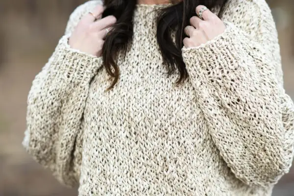 Primo piano di un maglione a maglia color crema con filato a trama aperta. Le mani della donna, adornate con anelli, giocano con i suoi capelli scuri.