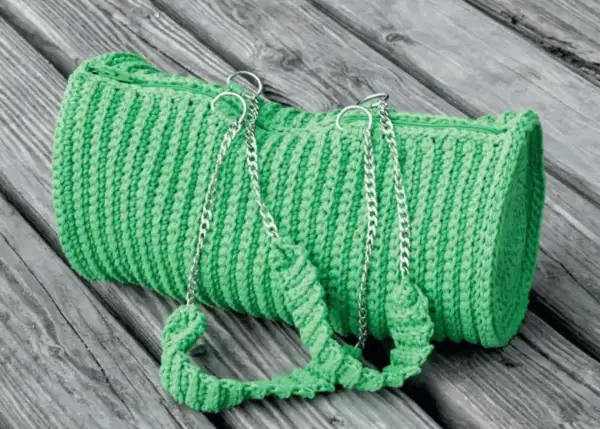 Borsa verde brillante lavorata all'uncinetto doppia punta, con manici e catene argentate, adagiata su assi di legno.