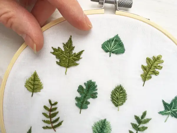 Mani che tengono un telaio da ricamo, con diverse piccole foglie ricamate su tessuto bianco. Un'arte delicata per ricamare foglie piccole.