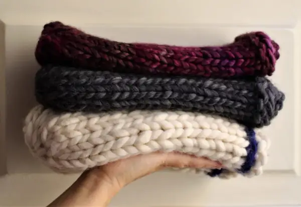 Tre berretti in lana ai ferri, uno bordeaux, uno grigio e uno bianco con striscia blu, impilati su una mano.