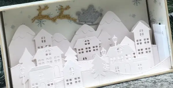 Sagome di un villaggio di Natale da ritagliare, con renne e slitta di Babbo Natale che volano sopra le casette innevate.