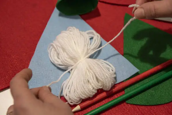 Mani che preparano lavoretti di Natale con la lana facili: gomitolo bianco, feltro colorato e matite.