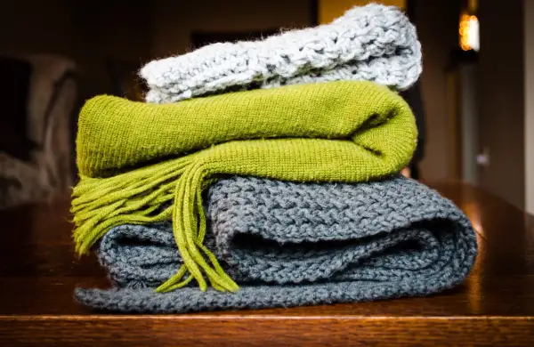Pile di sciarpe ai ferri: una grigia, una verde con frange e una grigio chiaro in cima. Quanti punti per una sciarpa ai ferri? Dipende dalla lana e dalla maglia!