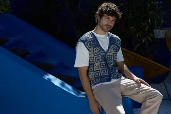 Uomo con riccio gilet senza maniche all'uncinetto blu e beige, seduto su una struttura blu.